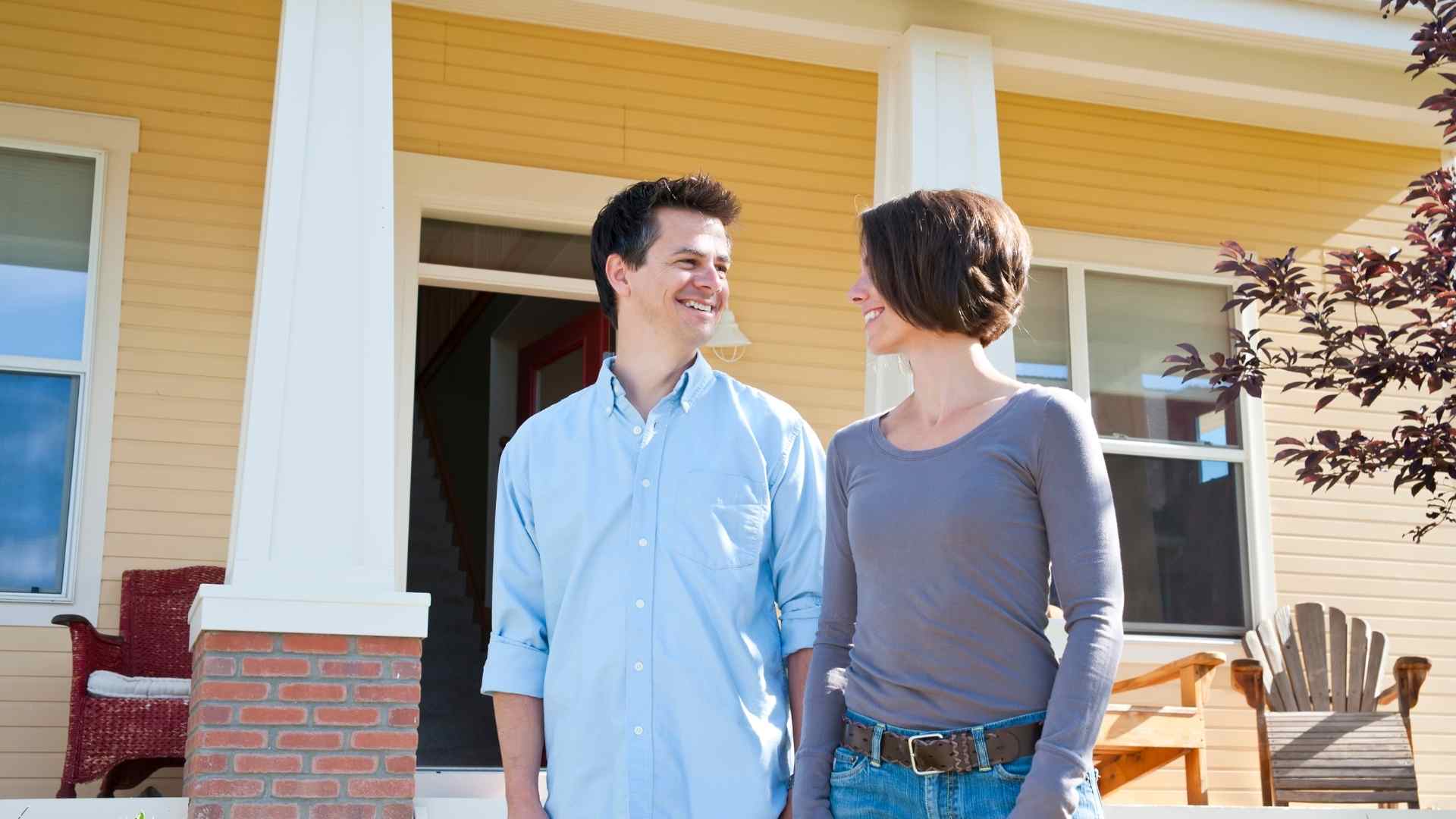 a smiling couple looking at each other and holding hands in front of yellow home