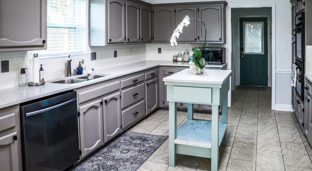 A renovated kitchen in an older home with painted gray cabinets, marble countertops, a small portable island and a tiled floor